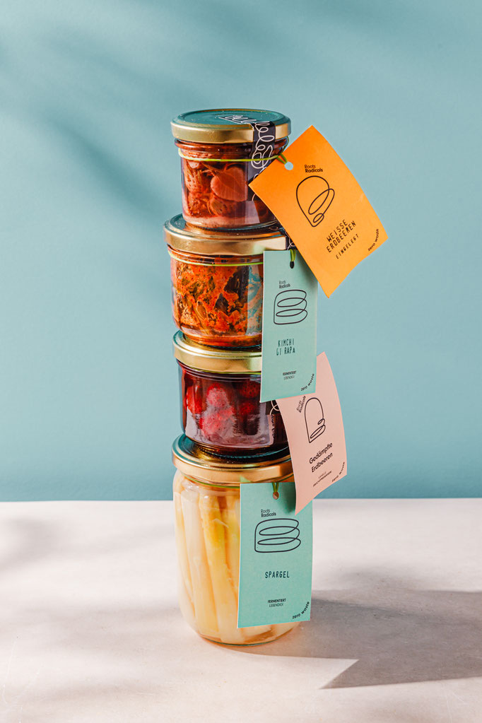 Stapel aus vier farbenfrohen Gläsern mit fermentierten Lebensmitteln vor pastellblauem Hintergrund. Modernes Branding und Produktfotografie. Kooperation mit Zero Waste Food Company Roots Radicals aus Berlin. Styling, Fotogrfie, Rezeptentwicklung.