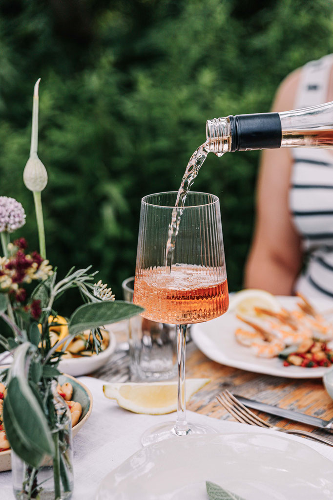 Roséwein wird an festlich gedecktem Tisch eingeschenkt – sommerliche Gartenszene mit Fokus auf Genuss, regionale Zutaten und geselliges Essen mit Garnelen