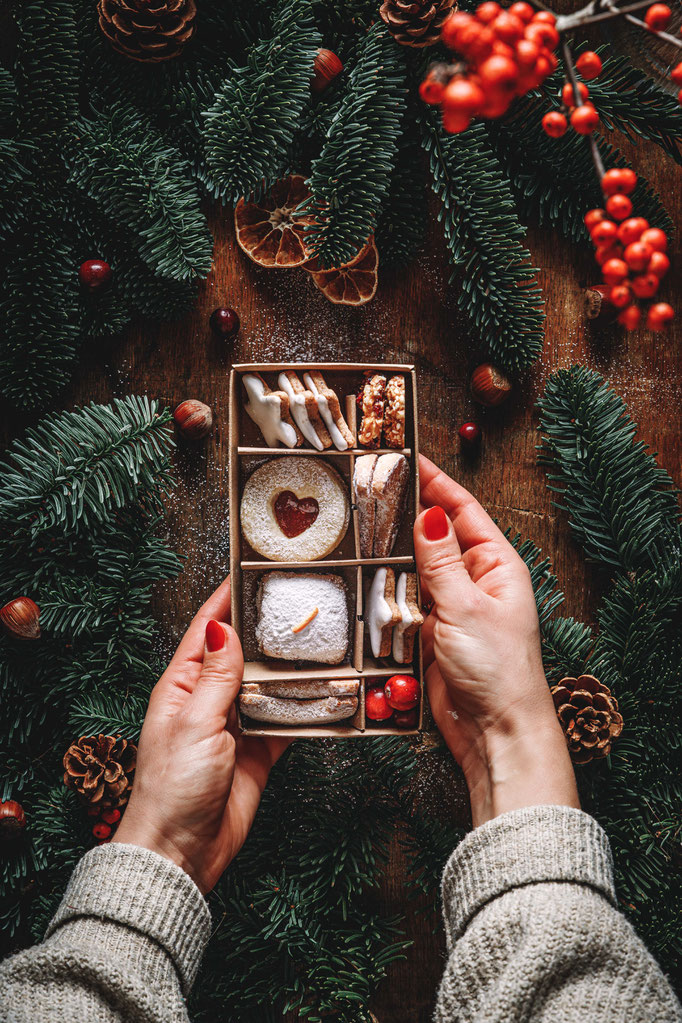 Hände halten eine kleine Geschenkschachtel mit verschiedenen Weihnachtsplätzchen, dekoriert mit Tannenzweigen, Zapfen und roten Beeren.“