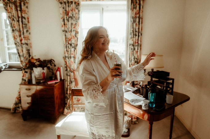 Braut lacht beim Getting Ready mit einem Getränk in der Hand