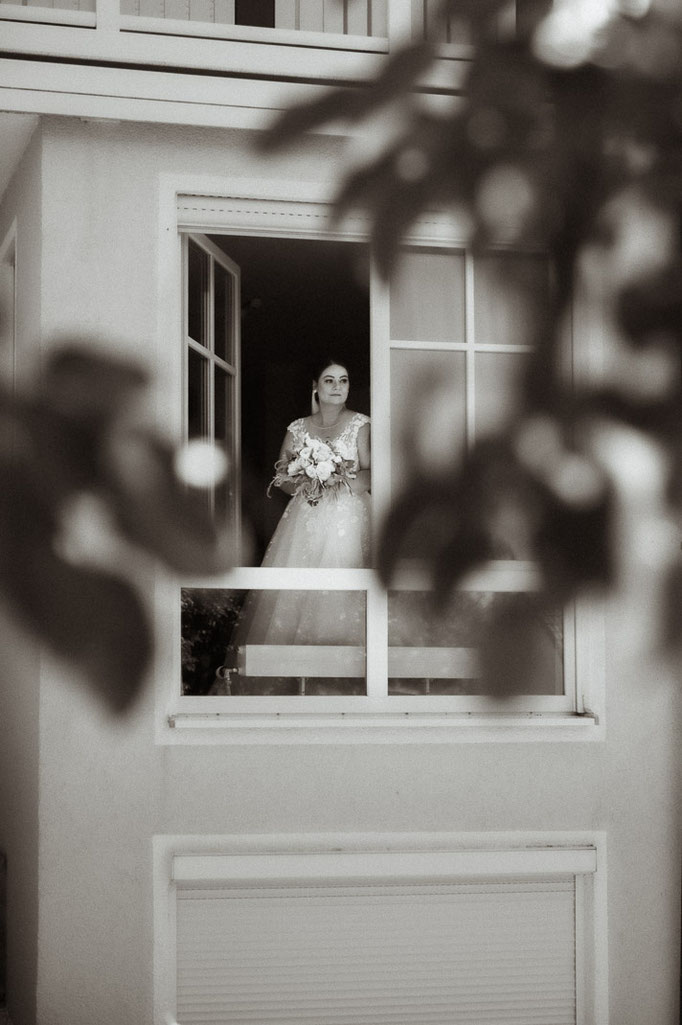 Die fertig gestylte Braut blickt aus dem Fenster in die Ferne, entstanden beim Getting Ready, fotografiert von uns als Hochzeitsfotografen