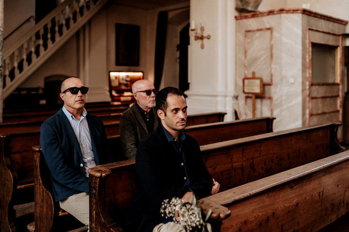 Hochzeitsfotograf in der St. Sixtus Kirche am Schliersee. Die Hochzeitsgesellschaft hört der Predigt gespannt zu, hinten sitzen zwei Männer mit Sonnenbrillen.