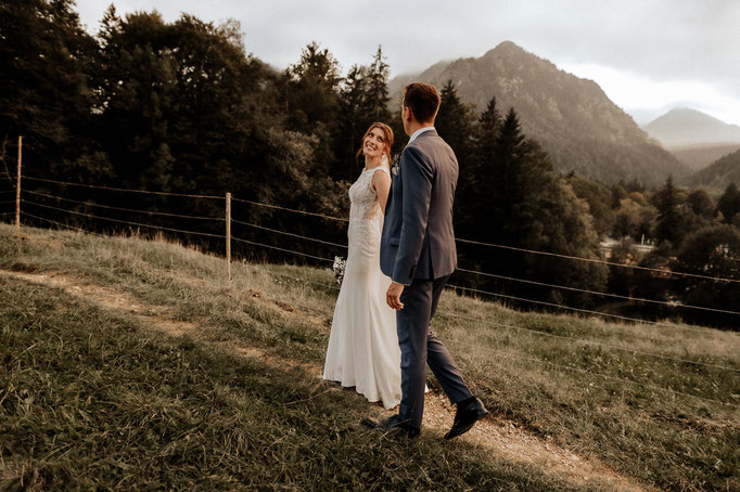 Die Braut sieht verliebt zu ihrem Bräutigam zurück, im Hintergrund sind Berge zu sehen. Brautpaarfotos beim Wasmeier Freilichtmuseum am Schliersee. 