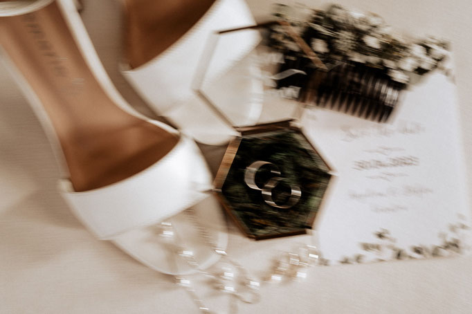 Brautschuhe, Brautschmuck, Eheringe in einer Glasschatulle, Haarkamm und Hochzeitspapeterie als verschwommenes Stillleben