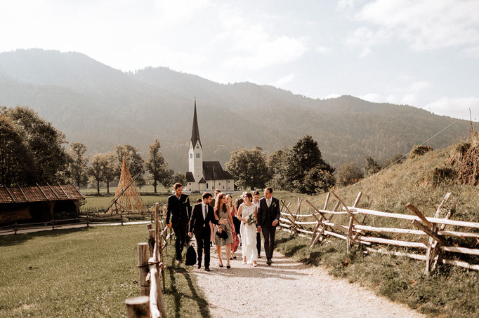 Hochzeitsfotograf am Schliersee, die Hochzeitsgesellschaft geht im Wasmeier Freilichtmuseum zu ihrer Feierlocation. Im Hintergrund ist eine Kirche und Berge zu sehen.