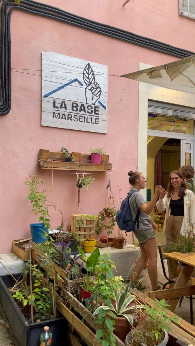 Bildbeschreibung: An der Wand hängt ein Schild mit dem Logo von "La Base Marseille". Davor Pflanzen in Blumentöpfen und Kisten und ein paar Personen.