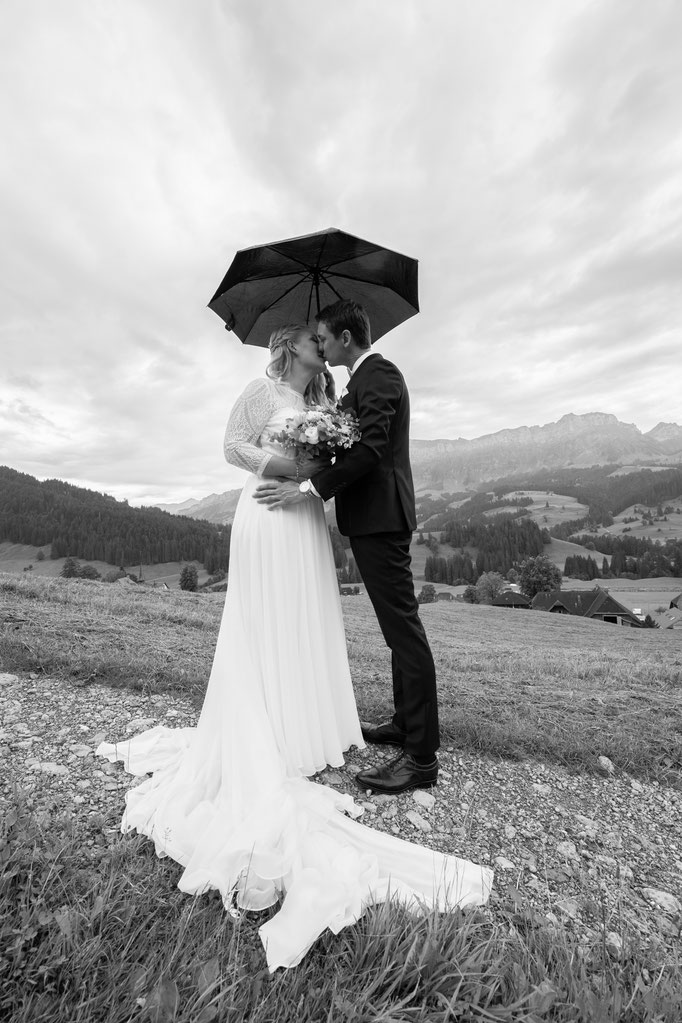 Schwarz-weiss Fotografie eines Brautpaars, das sich auf einer Wiese unter einem Regenschirm küsst
