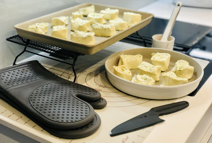Hier siehst du meine vorbereiteten Käse-Schnecken und alle Produkte die ich hierfür verwendet habe: Teigunterlage, Nylonmesser, Silikonpinsel, Silikonhandschuhe PacksAn, Kuchengitter, großer Ofenzauberer und die runde Stoneware Leila