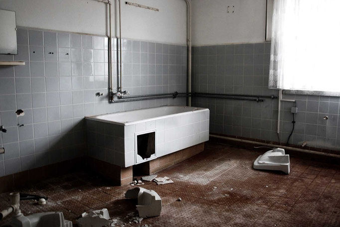 Ein verlassnes Badezimmer mit blauen Fliesen an den Wänden und einer Badewanne. Auf dem Boden liegen zerschlagene Waschbecken.