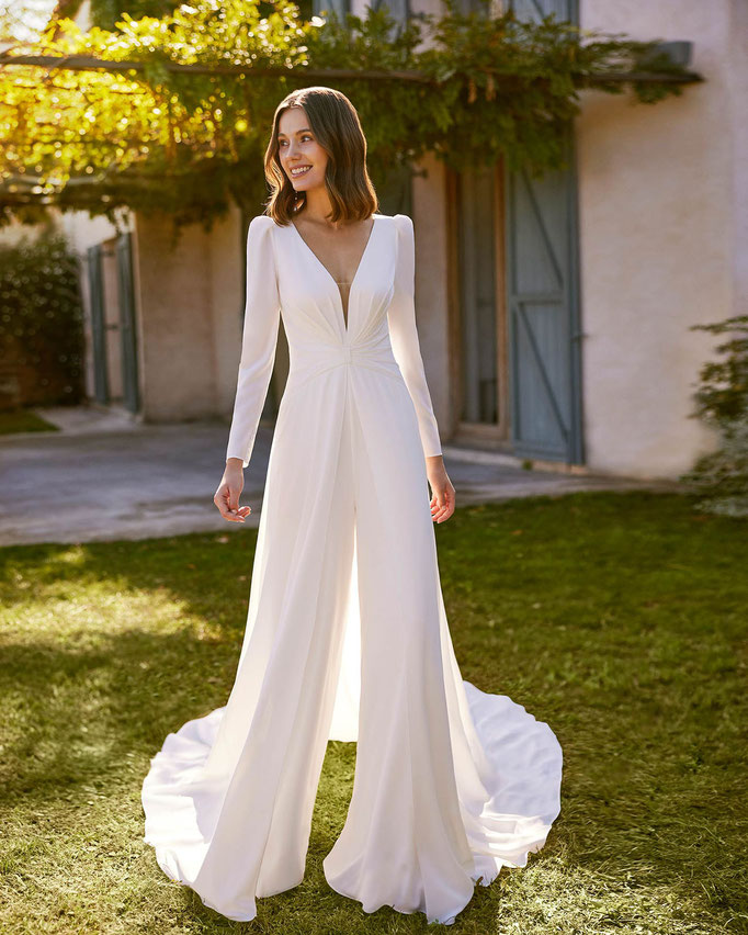 Junge Frau in einem schicken Vorgarten trägt das Jumpsuit-Brautkleid Naelia aus der Alma Novia Kollektion 2026