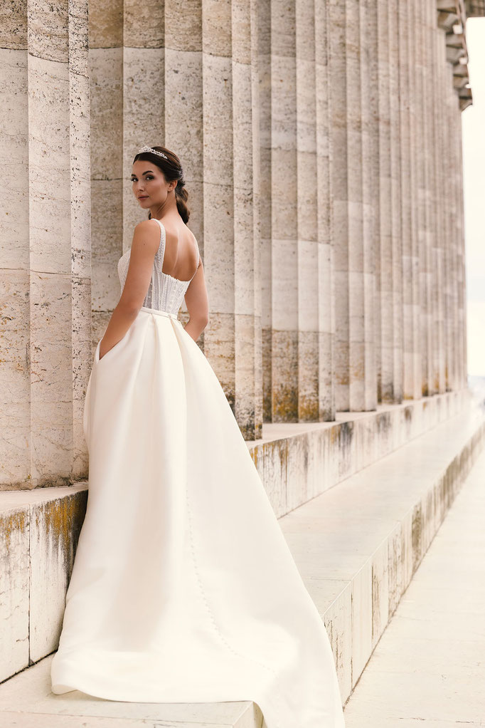 Junge Frau neben Steinsäulen trägt das Brautkleid Catharina aus der Amo & Luv Kollektion
