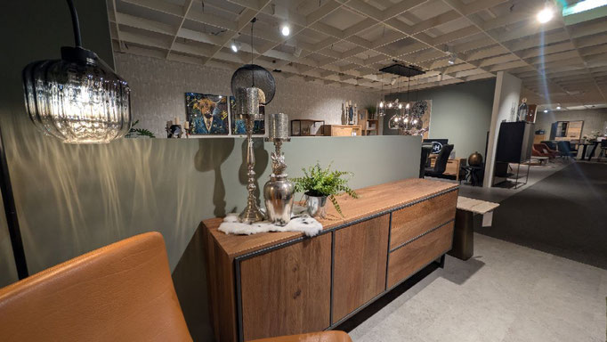 Holz-Sideboard mit zwei silbernen Kerzenhaltern, Farn und braunem Sessel in einem Möbelhaus.