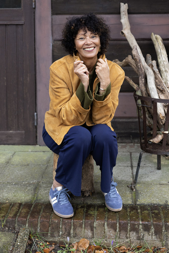 Yolanda zittend op een boomstam voor een huisje op landgoed Duinlust. Tijdens de Fotoshoot Dag gefotografeerd door Stefan van Ruijven Fotografie om haar portfolio een update te geven. Ze staat met een commerciële lach op de foto en een casual outfit