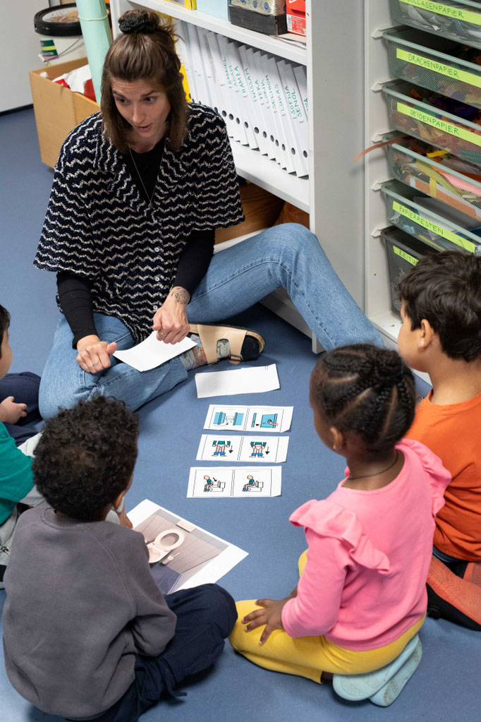 Lehrerin_sitzt_mit_kleiner_Gruppe_von_Kindern_am_Boden_erklaert_wie_WC_funktioniert