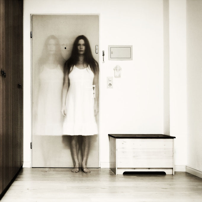 Manuela Deigert Bildsprache, Langzeitbelichtung eines Selbstportraits in doppelter Ansicht, stehend in einem Raum vor einer Tür, seitlich jeweils eine Holzkiste und Schrank