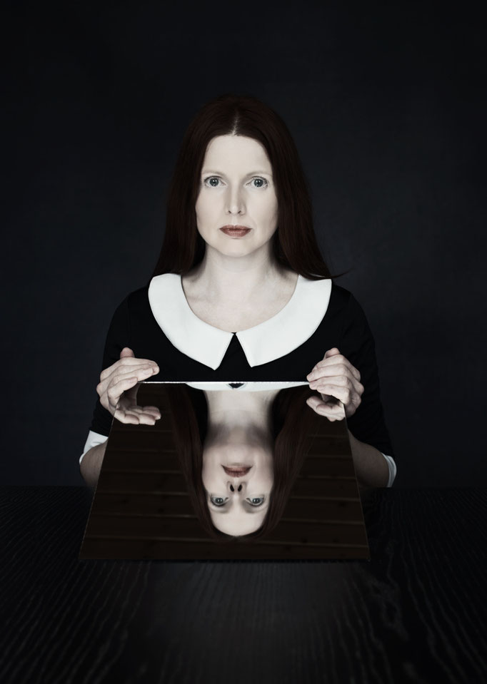 Manuela Deigert Bildsprache Selbstportrait an einem Tisch und einen Spiegel haltend, auf dem mein Gesicht reflektiert wird