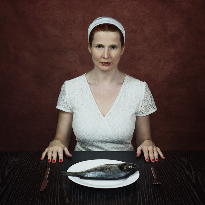 Manuela Deigert Projekte Friday is fishday, farbiges Selbstportrait an einem Tisch sitzend mit einem Teller auf dem eine rohe Dorade liegt