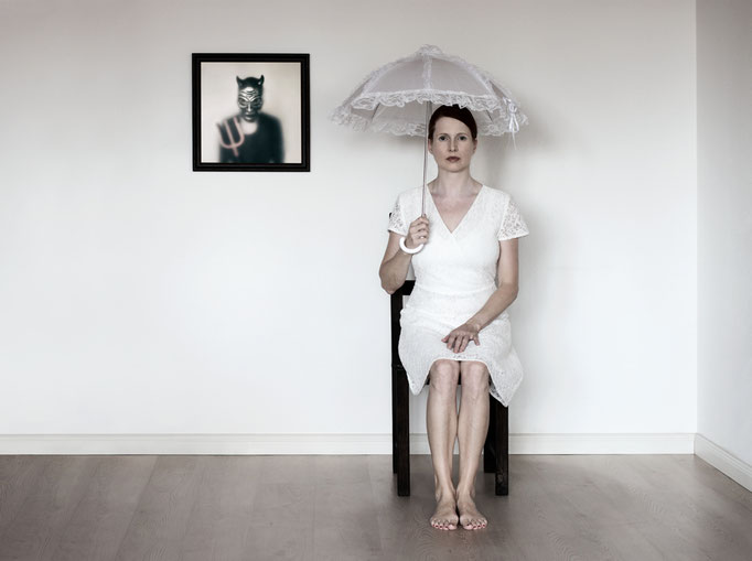 Manuela Deigert Bildsprache Selbstportrait auf einem Stuhl sitzend mit aufgespannten Schirm und einem Bild vom Teufel an der Wand