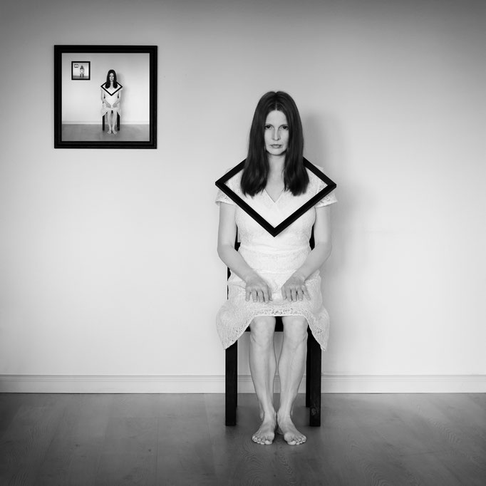 Manuela Deigert Bildsprache Selbstportrait im sitzen auf einem Stuhl mit Rahmen um den Hals und einen Bilderrahmen daneben mit surrealer Spiegelung des Abbildes