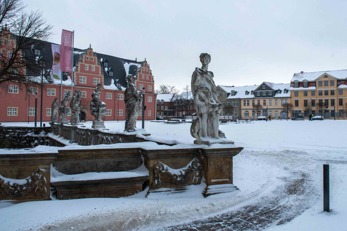 Ein großer schneebedeckter Platz an dem mehrere barocke Gebäude stehen. Auf einem Geländer stehen mehrere aus weißem Stein gefertigte Figuren.