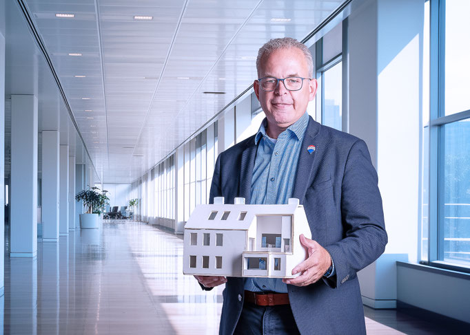 Portrait eines Herren mit Modell eines Hauses in den Händen vor virtuellem Büro-Hintergrund