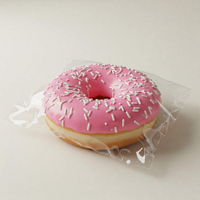 En una mesa blanca con un fondo blanco hay una dona de chocolate rosa con granillo de chocolate blanco. La dona está empacada en una bolsa de celofán transparente de medida 14x14. La bolsa tiene adhesivo lo que permite cerrar la bolsa