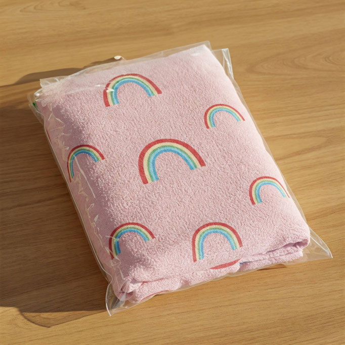 Encima de un mesa de madera de color maple hay una cobija de color rosa pastel con varias imágenes de arcoíris. La cobija está empacada en una bolsa de celofán con adhesivo de 20x30 para poder abrir y cerrar la bolsa.