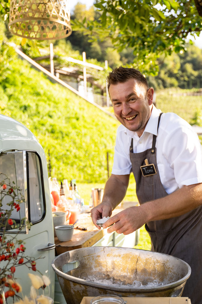 A smiling barman in an apron is preparing drinks with ice outside next to a small white car, surrounded by summer greenery and warm sunlight.