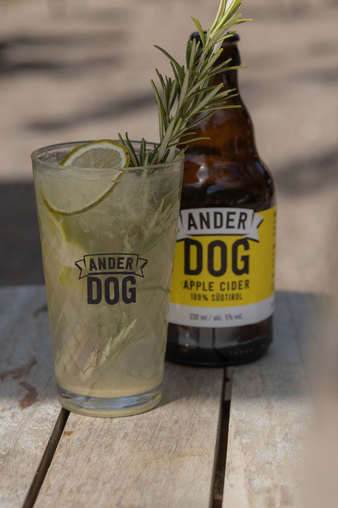 A refreshing cocktail with lime slices, ice and a sprig of rosemary in a glass, placed in front of a bottle of “Ander Dog” apple cider on a wooden table outdoors.