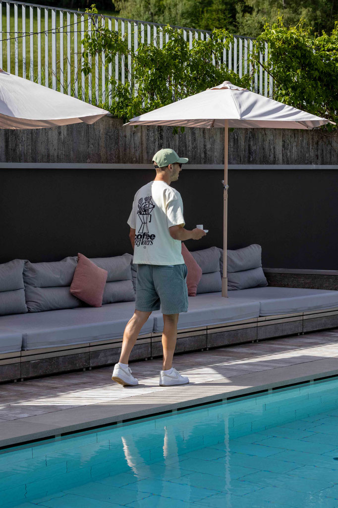 Gast spaziert mit einer Tasse Kaffee am Außenpool des Boutiquehotel PETRUS in Reischach-Bruneck • Südtirol entlang und genießt die entspannte Atmosphäre im Garten.