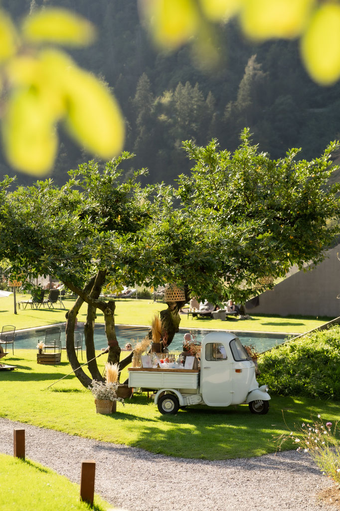 Kleiner weißer Piaggio-Dreiradwagen mit dekorierter Bar-Aufbauten steht auf einer sonnigen Wiese unter Bäumen nahe eines Pools und Spa-Geländes in alpiner Naturkulisse.