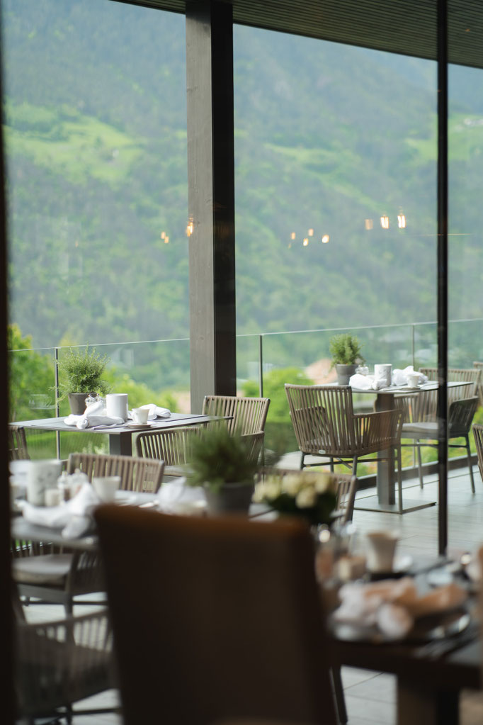 A view from the restaurant at the Hotel Mair in Living, Partschins, near Merano in South Tyrol, looking out onto the sunny terrace with its set tables and the surrounding mountain landscape. The generous glass fronts create a bright, open and inviting atm