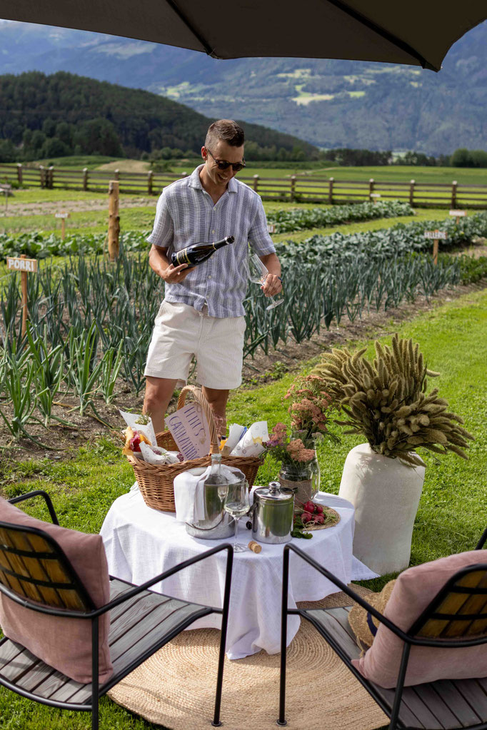Gast genießt ein Picknick mit regionalen Spezialitäten und einem Glas Wein im Garten des Boutiquehotel PETRUS in Reischach-Bruneck • Südtirol, umgeben von Gemüsefeldern und der beeindruckenden Südtiroler Berglandschaft.