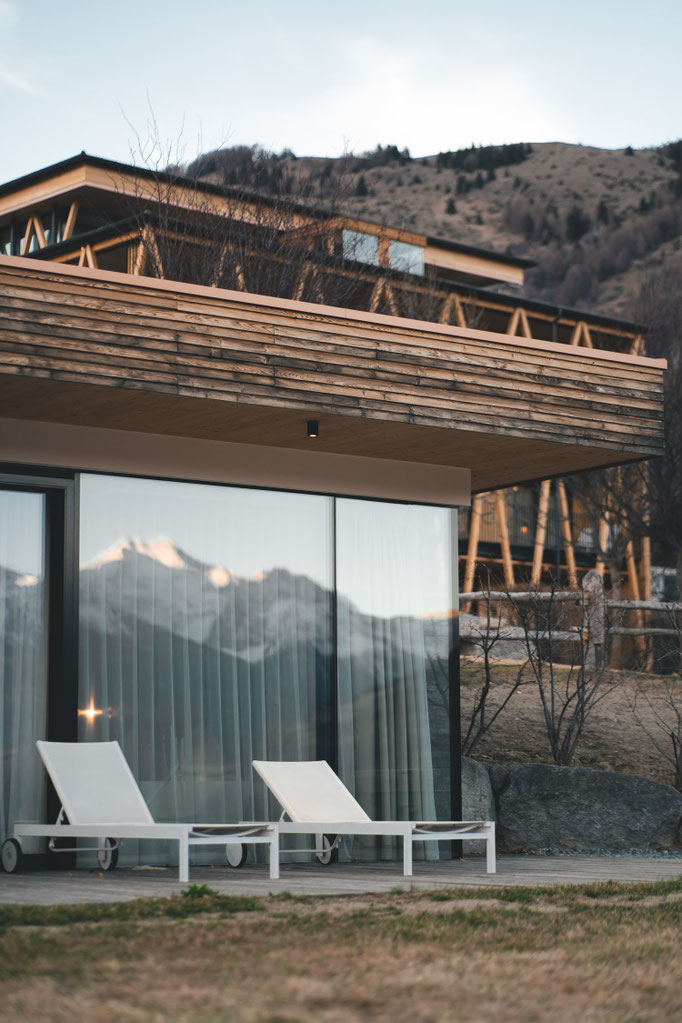 A modern hotel terrace with sun loungers, with the mountains reflected in the glass façade.