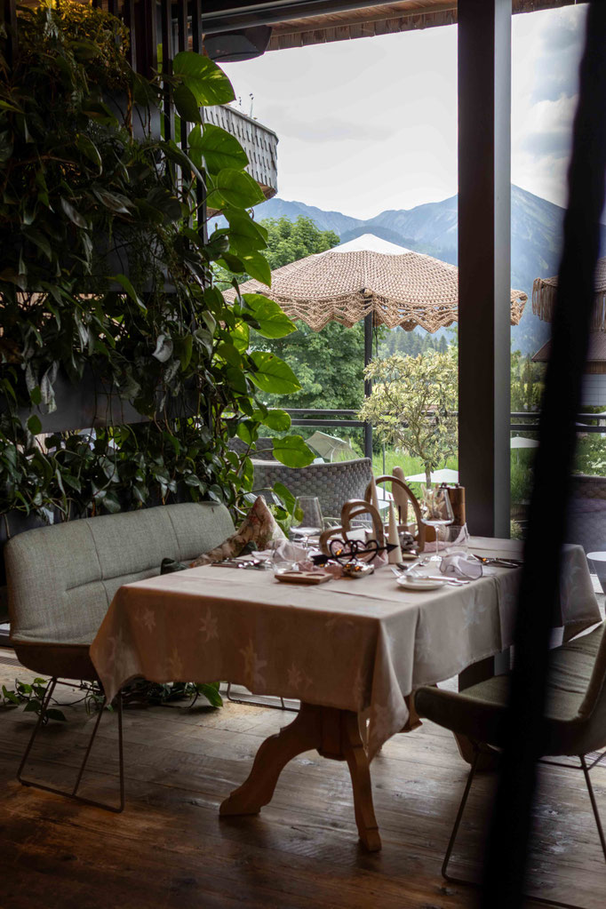 Stilvoll gedeckter Tisch im Restaurant des DER ENGEL im Tannheimertal mit Blick ins Grüne und auf die Bergwelt. Natürliche Materialien, üppige Pflanzen und die gemütliche Atmosphäre machen jeden Genussmoment zu einem besonderen Erlebnis.