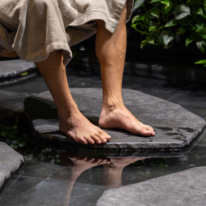 Eine Person mit einem hellen Handtuch um den Körper steht barfuß auf großen Trittsteinen in einem flachen Wasserbecken. Das Wasser spiegelt die Füße wider, während üppige grüne Pflanzen im Hintergrund eine ruhige Spa-Atmosphäre schaffen.