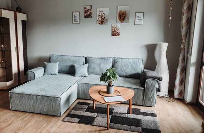 Modernes Wohnzimmer mit großer, hellgrauer Eckcouch, rundem Holztisch und stilvollem Teppich. Wandbilder in Naturtönen und eine große Stehlampe runden die gemütliche Atmosphäre in unserer Ferienwohnung in Winterberg-Niedersfeld ab.