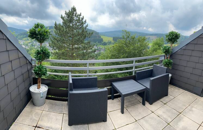 Gemütlicher Balkon mit Sitzmöbeln und Tisch in unserer Ferienwohnung in Winterberg-Niedersfeld. Umgeben von grünen Bäumen und Hügeln lädt der Ausblick ins Sauerland zur Entspannung und Erholung ein – perfekt für eine Auszeit in der Natur.