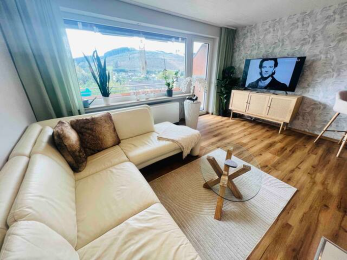 Helles Wohnzimmer mit großer Fensterfront und Blick ins Grüne in unserer Ferienwohnung in Winterberg-Niedersfeld. Ausgestattet mit bequemer Eckcouch, TV, stilvollem Teppich und dekorativen Pflanzen – ideal zum Entspannen und Genießen