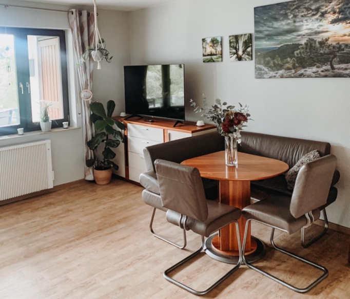 Heller Essbereich in unserer Ferienwohnung in Winterberg-Niedersfeld mit rundem Holztisch, bequemer Eckbank und Flachbild-TV. Große Fenster, Pflanzen und stilvolle Wandbilder sorgen für eine gemütliche und wohnliche Atmosphäre.