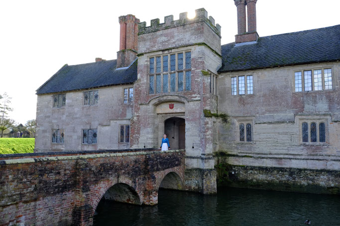 Ich stehe auf der Brücke des Anwesens im Haupttor in einem langen weißen Kleid und Biedermeier-Spencer. This image shows Baddesley Clinton, a historic home, with a bridge on which I stand in a white Regency Dress and blue Spencer