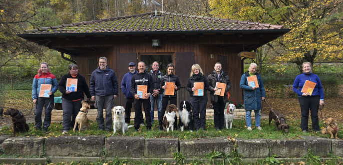 Gruppe von Menschen steht vor einem Holzhaus, jeder hält eine orangefarbene Urkunde, mehrere Hunde sitzen oder stehen vor ihnen auf einer Wiese mit herbstlichen Bäumen im Hintergrund.