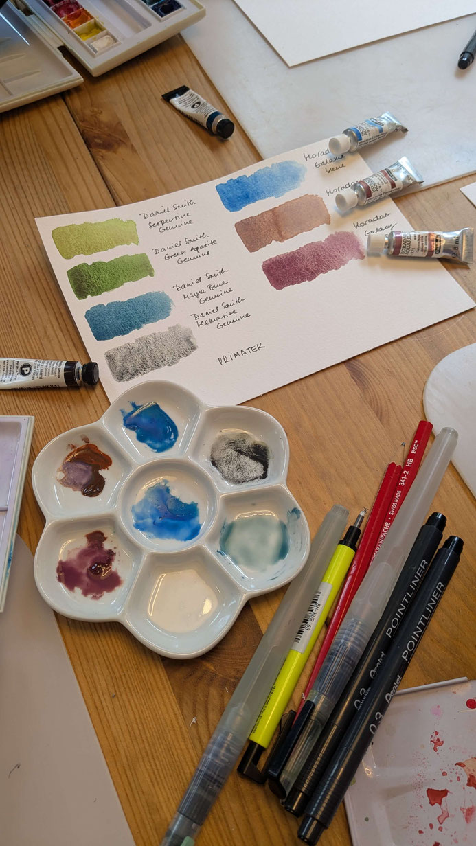 Une table de peinture à l'aquarelle à grannulation avec des pinceaux, des crayons, des tubes de peinture et une palette de mélange. Une feuille montre des échantillons de couleurs annotés. Des stylos et des crayons sont également visibles