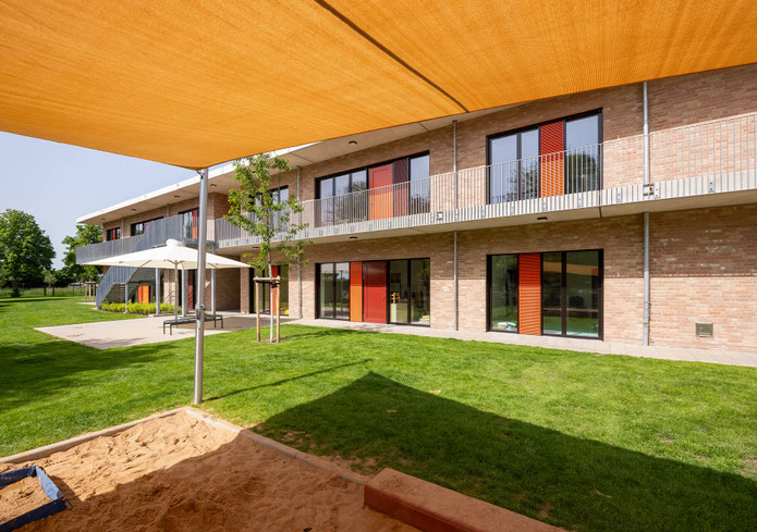 Neubau Kita Bergheim-Ahe Ansicht Kinderperspektive aus dem beschatteten Sandkasten auf das Gebäude