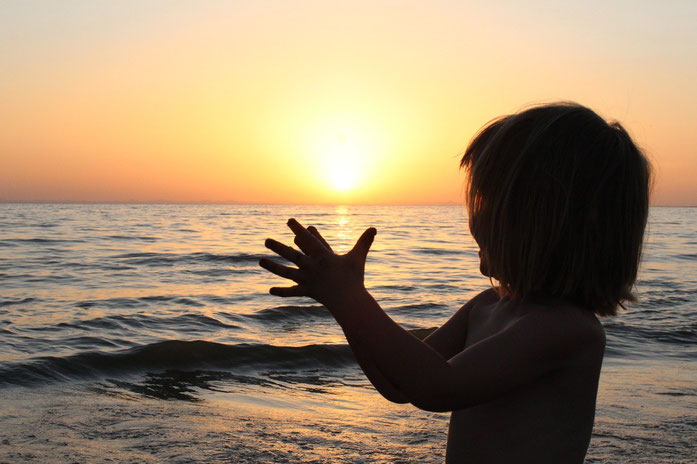 Photographie d'une petite fille joignant les mains devant un coucher de soleil sur la mer