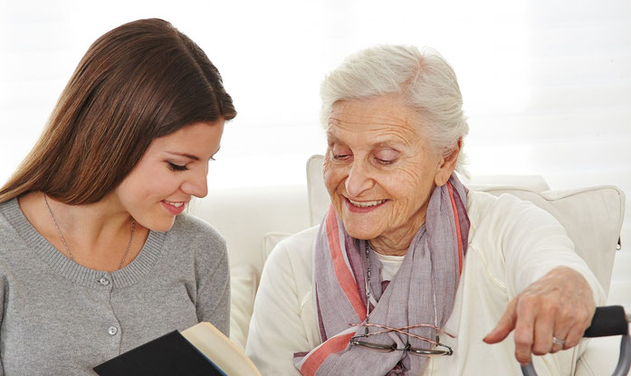 Seniorenbegleitung. Eine junge Frau leistet einer Seniorin Gesellschaft und liest vor.