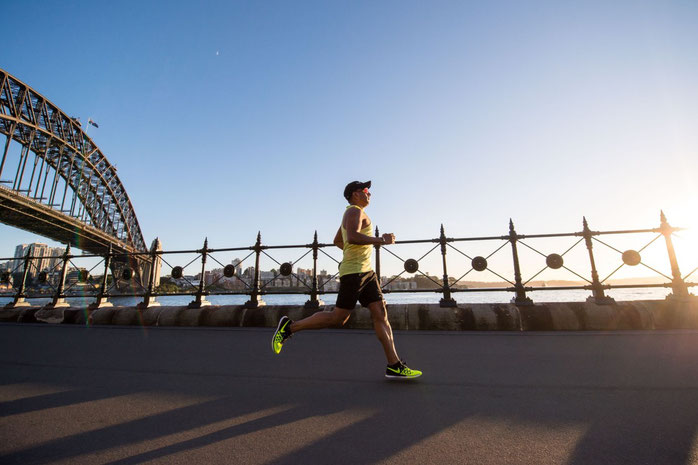 Ein Sportler löuft auf einer Brücke am Wasser einen Marathon. Im Hintergrund sieht man das Wasser und die Sonne.