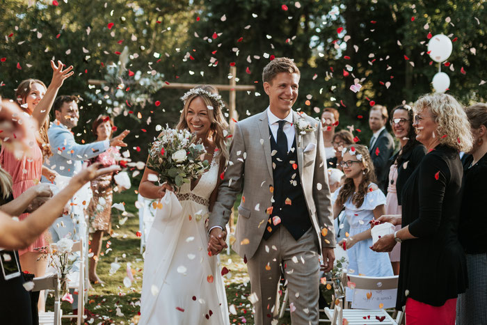 Das Hochzeitspaar läuft durch einen Regen aus Blüten, für den die Gäste sorgen. Ein Durchgang und Bäume im Hintergrund