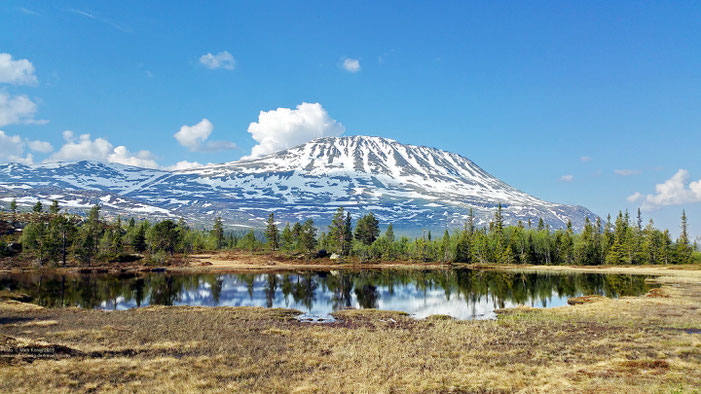 Photo of snow-capped mountain Gaustatoppen with blue sky, a partly mirrorig lake in the foreground