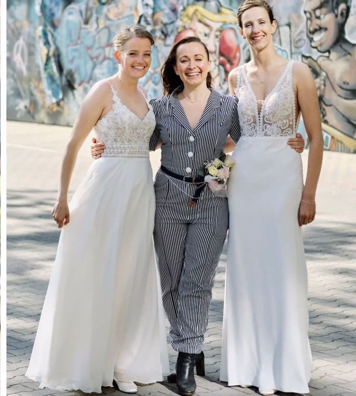 Constanze Marienfeld lächelt stolz, im Arm hält sie ihr Brautpaar, zwei Frauen, beide im Brautkleid